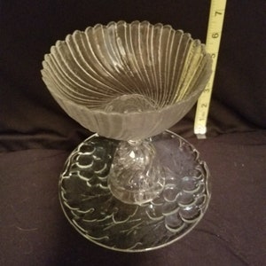 May include: A clear glass pedestal bowl with a fluted design and a decorative base. The bowl is on a clear glass plate with a textured design.