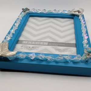 May include: A turquoise picture frame with a decorative border of clear, faceted beads. Silver-colored cowboy boot, horseshoe, and turquoise accents adorn the corners. The frame holds a 4-inch x 6-inch (10.16 cm x 15.24 cm) photo.