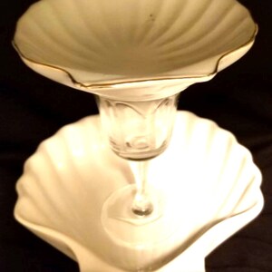 May include: A white ceramic two-tiered serving dish shaped like seashells. The top tier is a shallow bowl with a gold rim. The bottom tier is a larger bowl with a scalloped edge. A clear glass pedestal connects the two tiers.