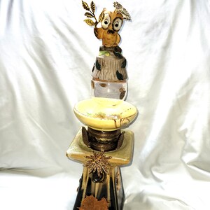 May include: A decorative owl figurine with a gold leaf branch and a flower crown sits atop a multi-tiered stand. The stand features a yellow dish, a square base, and a black and gold color scheme with leaf accents. The owl is brown and tan.
