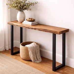 Può includere: Una consolle in legno con piano a bordo vivo e gambe in metallo nero. Un vaso bianco con verde, libri e un cestino intrecciato con una coperta sono sopra o sotto il tavolo. La stanza ha una palette di colori neutri.
