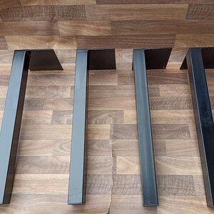 Könnte beinhalten: Vier schwarze Metalltischbeine mit quadratischem Ober- und Unterteil. Die Beine stehen auf einem Holzboden.