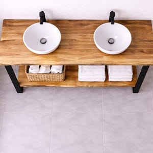Peut inclure: Meuble-lavabo en bois avec deux lavabos ronds blancs et des robinets noirs. Le meuble a une étagère inférieure avec un panier en osier et des serviettes blanches pliées. La base est en métal noir, et le comptoir est en bois naturel.