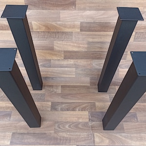 Könnte beinhalten: Vier schwarze Metall-Tischbeine mit quadratischen Montageplatten. Die Beine sind rechteckig und verjüngen sich leicht nach unten. Die Beine sind aus Metall und schwarz lackiert.