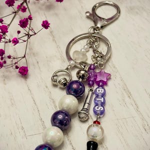 May include: A silver keychain featuring a metal ring and lobster clasp. It includes dangling charms: a silver headphone charm, a silver microphone charm, a purple star bead, and beads spelling BTS. The design incorporates purple and white pearls.