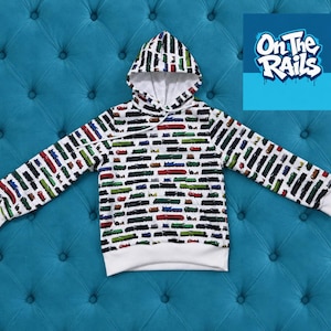 Könnte beinhalten: Ein weißer Kapuzenpullover mit einem sich wiederholenden Muster aus bunten Spielzeugeisenbahnen in Rot, Grün, Blau und Schwarz. Der Hoodie hat lange Ärmel, einen weißen Kordelzug sowie weiße gerippte Bündchen und einen Saum. Der Text On The Rails ist sichtbar.