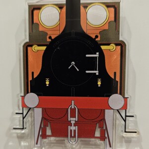May include: A colorful train-shaped acrylic charm. The design features a black engine with a red front, orange accents, and a yellow top. The charm is a fun accessory for train enthusiasts.