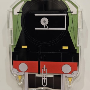 May include: A clear acrylic train-shaped ornament. The design features a black locomotive with green and red accents. The front of the train is black with a white gauge and a red bumper. The ornament is a cut-out shape.