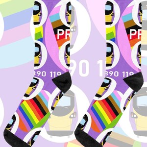 Op de afbeelding: Een paar paarse sokken met een treindesign, regenboogstrepen en de tekst "PR". De sokken hebben zwarte hielen en tenen. De tekst "On The Rails" staat in een blauwe rechthoek.