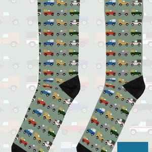 May include: A pair of sage green crew socks featuring a repeating pattern of small, colourful off-road vehicles in red, blue, yellow, and green. The socks have solid black heels and toes. A blue square logo with the text On The Rails is visible.