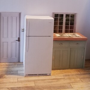 Puede incluir: Un refrigerador blanco con un compartimento congelador en la parte superior, un armario verde con dos puertas y dos cajones, y una ventana con vista a los árboles. El refrigerador está en una cocina con suelo de baldosas de imitación de madera.