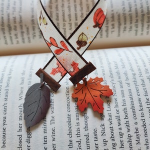 May include: A white ribbon bookmark with a fall theme. The ribbon has a pattern of orange and brown leaves, acorns, and pumpkins. The bookmark is attached to two fall-colored leaf charms.