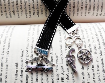 May include: Black bookmark with white stitching and silver charms. Charms include a car with wings, a revolver, a pentagram, and handcuffs. The bookmark is on a book with printed text.