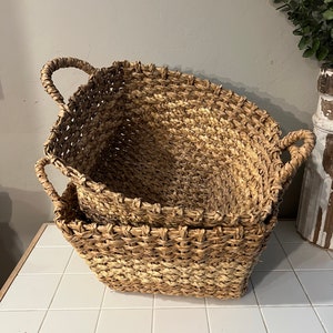 May include: Two woven baskets with handles, one nested inside the other. The baskets are made of a light brown, natural material and have a rectangular shape. They are sitting on a white tiled surface.