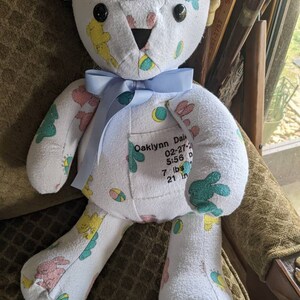 Pode incluir: Um urso de peluche branco com uma fita azul em volta do pesco&ccedil;o. O urso &eacute; feito de roupas de beb&ecirc; e tem uma etiqueta costurada no peito que diz "Oaklynn Dale 02-27-20 5:56 p 7 lbs 21 in".