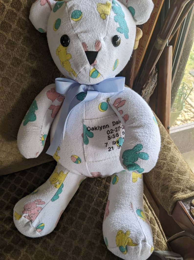 Pode incluir: Um urso de peluche branco com uma fita azul em volta do pesco&ccedil;o. O urso &eacute; feito de roupas de beb&ecirc; e tem uma etiqueta costurada no peito que diz "Oaklynn Dale 02-27-20 5:56 p 7 lbs 21 in".