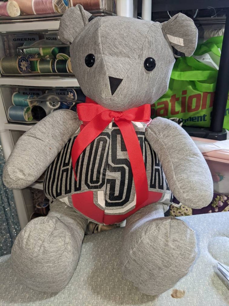 Pode incluir: Um urso de peluche cinza com um la&ccedil;o vermelho e uma camisa preta e branca com a palavra "HOSTA" impressa.