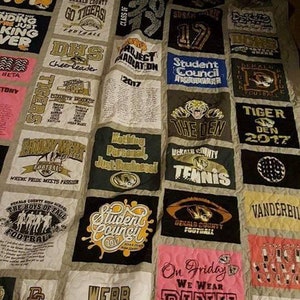 Pode incluir: Uma colcha feita de uma variedade de camisetas, com logotipos de escolas, equipes esportivas e outros designs. A colcha é feita em um estilo patchwork, com cada quadrado com um design diferente. A colcha é feita em uma variedade de cores, incluindo azul, verde, vermelho e branco.