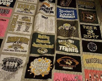 May include: A quilt made from a variety of t-shirts, featuring school logos, sports teams, and other designs. The quilt is made in a patchwork style, with each square featuring a different design. The quilt is made in a variety of colors, including blue, green, red, and white.