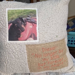 May include: A white throw pillow with a brown fabric patch that says "Diesel - No longer by my side but forever in my heart". The pillow also has a photo of a black and brown dachshund sitting on a red ATV.