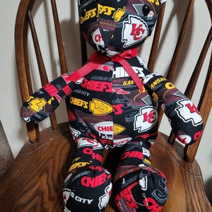 May include: A black and red plush teddy bear with a Kansas City Chiefs theme. The bear is wearing a red ribbon around its neck and has a white "KC" logo on its chest. The bear is sitting on a wooden chair.
