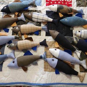 May include: A collection of handmade fabric fish in various colors and patterns, including blue, brown, and plaid. Each fish has fins and a tail, arranged on a quilted background. A creative and whimsical decorative item.