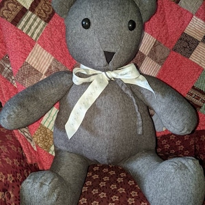 Pode incluir: Um urso de peluche de tecido cinza com olhos de bot&atilde;o pretos e uma fita branca ao redor do pesco&ccedil;o. O urso est&aacute; sentado em um edredom estampado de vermelho e marrom.