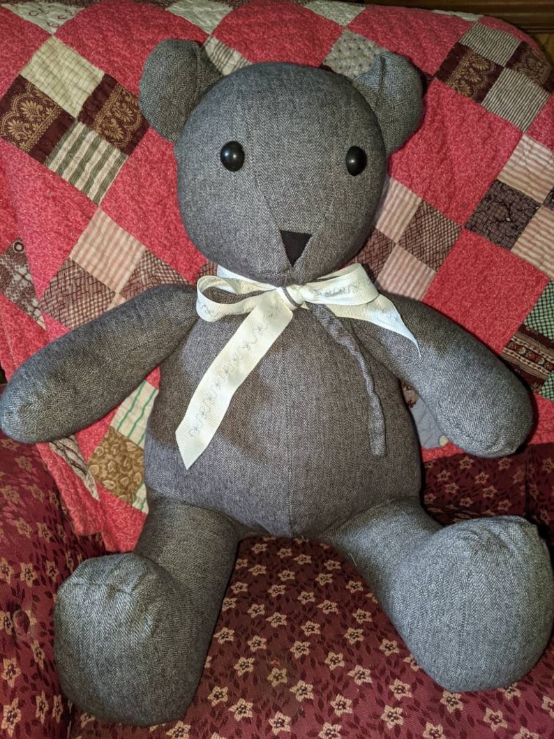 Pode incluir: Um urso de peluche de tecido cinza com olhos de bot&atilde;o pretos e uma fita branca ao redor do pesco&ccedil;o. O urso est&aacute; sentado em um edredom estampado de vermelho e marrom.