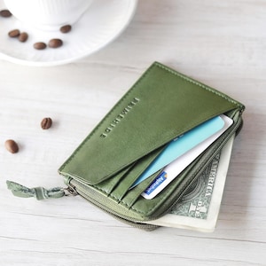 May include: Open olive green leather wallet with a zipper closure, containing credit cards and a dollar bill. The wallet has the brand name "REUMEHIDE" embossed on the front. A cup of coffee and a phone are in the background.