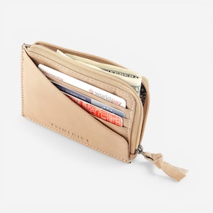 Op de afbeelding: Een bruinleren portemonnee met ritssluiting. De portemonnee is gedeeltelijk open en toont verschillende creditcards, contant geld en een klein vak. De ritssluiting heeft een klein leren lipje.