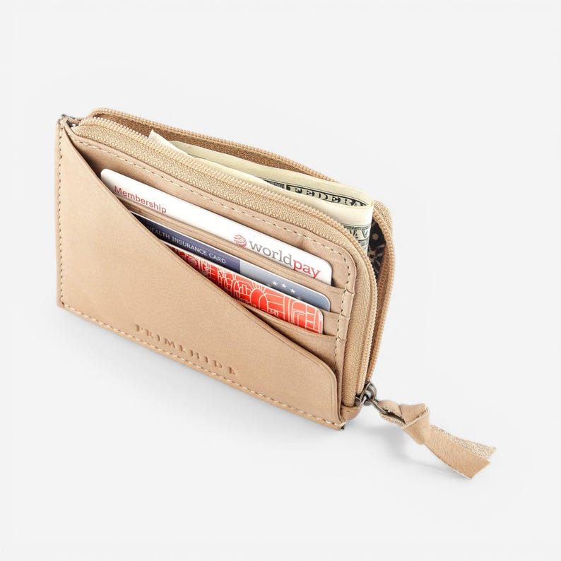 Op de afbeelding: Een bruinleren portemonnee met ritssluiting. De portemonnee is gedeeltelijk open en toont verschillende creditcards, contant geld en een klein vak. De ritssluiting heeft een klein leren lipje.