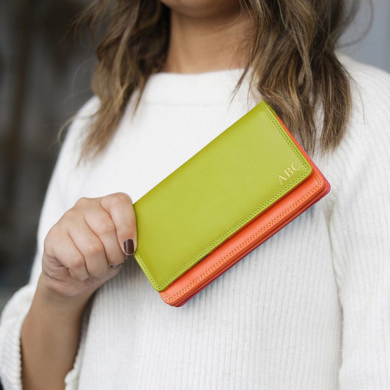 May include: A lime green and orange leather wallet with the initials "ABC" embossed in gold. The wallet is held by a person wearing a white sweater. The wallet is a long, rectangular shape.