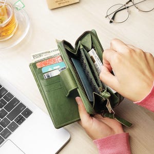 May include: A green leather wallet with multiple card slots and a zipped compartment. The wallet is open, revealing cards, cash, and other items. The wallet is on a light-coloured surface, with a laptop and a cup of tea in the background.