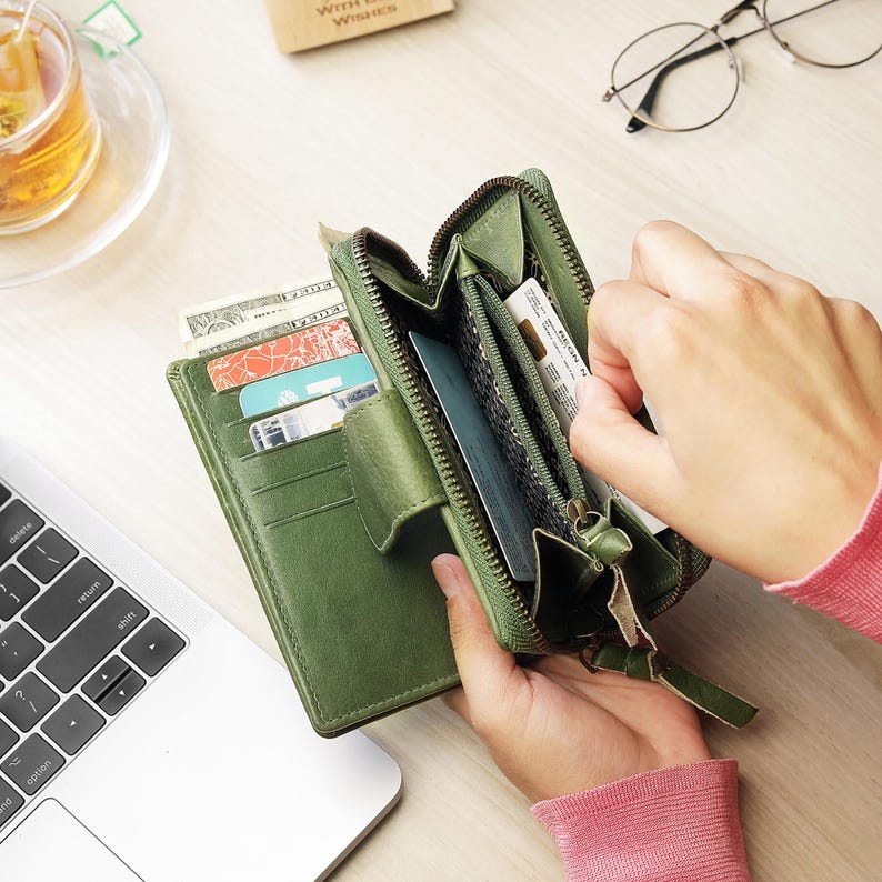 May include: A green leather wallet with multiple card slots and a zipped compartment. The wallet is open, revealing cards, cash, and other items. The wallet is on a light-coloured surface, with a laptop and a cup of tea in the background.