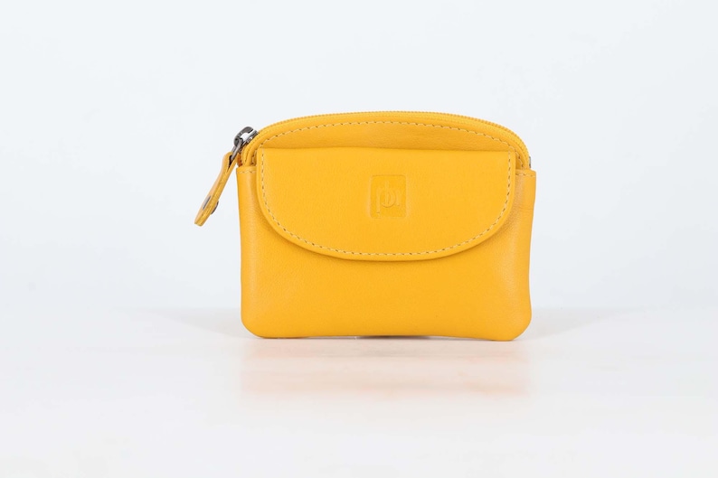 May include: A bright yellow coin purse with a smooth finish, featuring a top zipper closure and a front flap pocket with a small embossed square logo. The rectangular pouch has rounded corners and a matching yellow leather zipper pull tab.