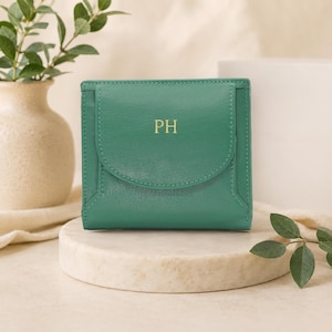 May include: A square, emerald green wallet featuring a rounded front flap closure and the gold-colored embossed initials PH centered on the front. The wallet has a smooth, matte texture and a structured design with visible stitching along the edges.