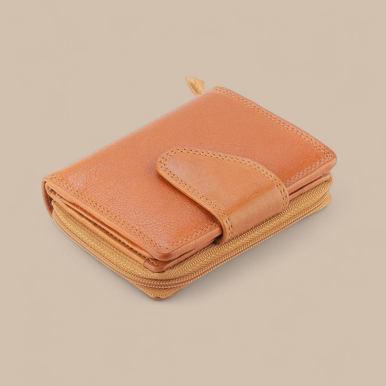 May include: A tan leather wallet featuring a textured surface, a central snap-button closure flap, and a wrap-around zipper along the perimeter. The wallet has a compact, rectangular shape with visible stitching along the edges and the closure flap.