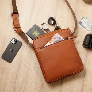 May include: A brown leather crossbody bag with a zipped pocket and a brown strap. The bag is open and contains a passport, cash, sunglasses, and a watch.
