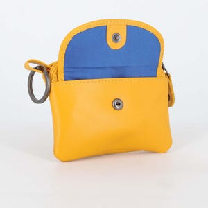 May include: A small yellow pouch with a rounded flap and a bright blue interior lining. The pouch features a silver-toned metal snap button closure on the front and a circular metal key ring attached to the side, providing a functional design.