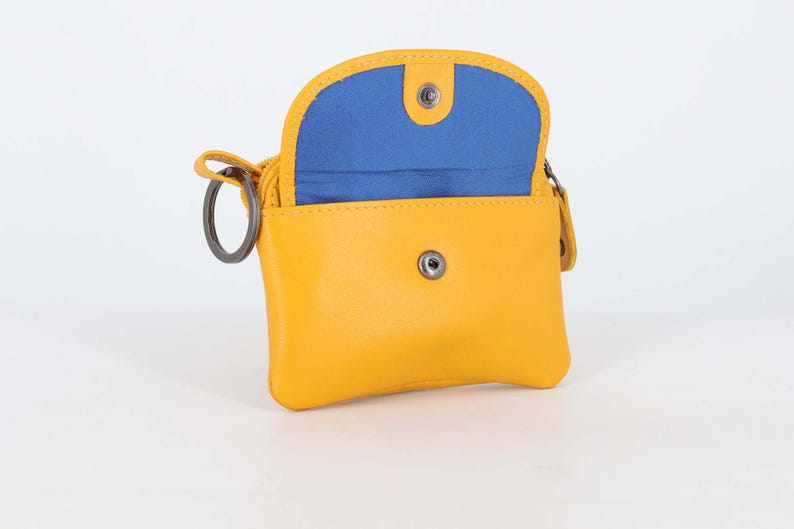 May include: A small yellow pouch with a rounded flap and a bright blue interior lining. The pouch features a silver-toned metal snap button closure on the front and a circular metal key ring attached to the side, providing a functional design.