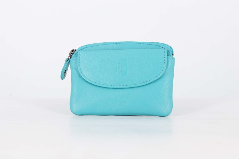 May include: A bright turquoise leather coin purse featuring a top zipper closure and a front flap pocket with a small embossed square logo. The compact accessory has a smooth texture and rounded edges, providing a simple design for carrying small items.