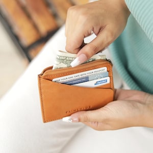 A tan leather wallet with a zip and a front pocket. The wallet is open, revealing a few dollar bills and some cards. "PRIMEHIDE" is printed on the front.