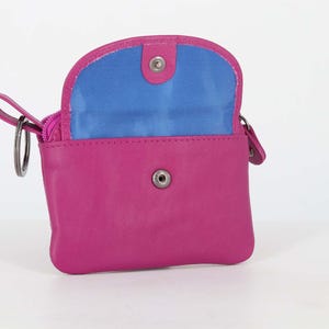 May include: A bright pink leather coin purse with a fold-over flap featuring a snap button closure. The interior lining is a vibrant blue. A metal key ring is attached to the side of the purse, which also includes a zippered compartment for storage.