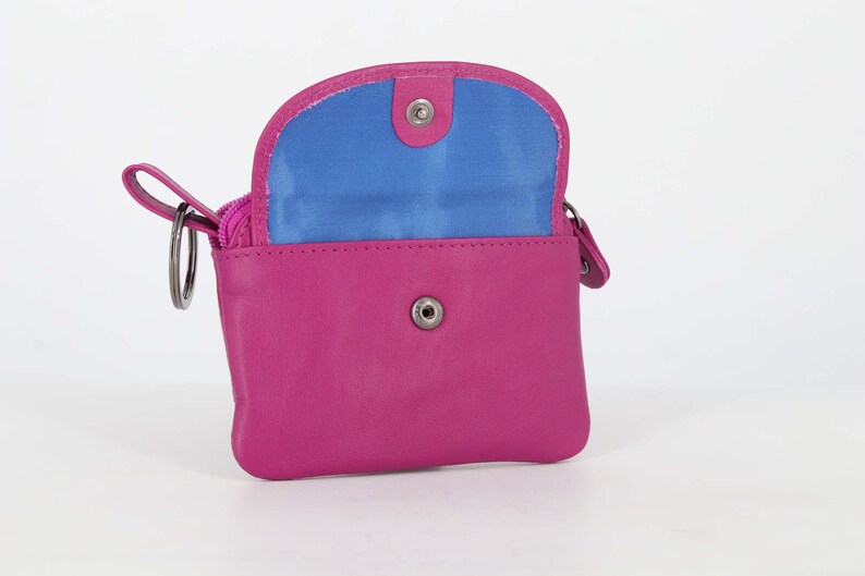 May include: A bright pink leather coin purse with a fold-over flap featuring a snap button closure. The interior lining is a vibrant blue. A metal key ring is attached to the side of the purse, which also includes a zippered compartment for storage.