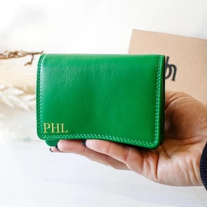 Pode incluir: Uma carteira de couro verde vibrante com as iniciais douradas "PHL" é segurada em uma mão. A carteira tem formato retangular com uma borda costurada. Uma sacola de papel marrom é visível ao fundo.