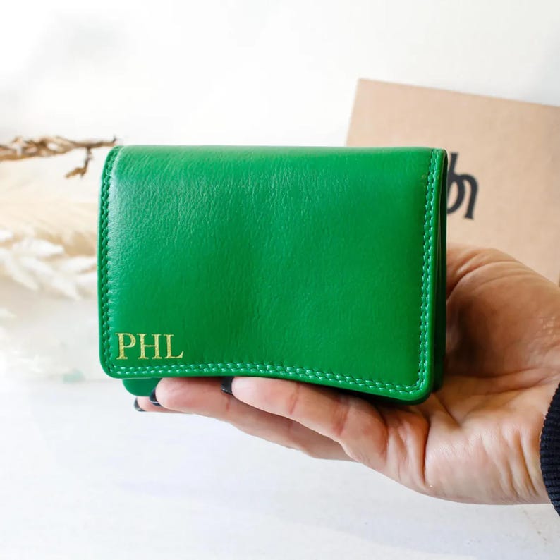 May include: A bright green leather wallet with gold initials "PHL" is held in a hand. The wallet is rectangular with a stitched edge. A brown paper bag is visible in the background.