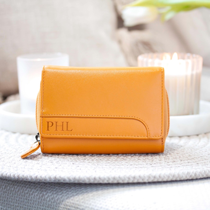 May include: A mustard yellow leather wallet featuring a fold-over flap with a curved stitched detail and the embossed letters PHL on the front. The wallet includes a side zip closure with a matching leather pull tab for secure storage.