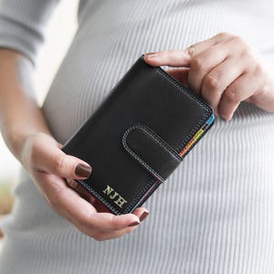 Może przedstawiać: Czarny skórzany portfel z białymi przeszyciami i zapięciem na zakładkę. Portfel ma tęczowe wnętrze, a na przodzie wytłoczone są inicjały "NJH". Portfel jest trzymany w dłoniach.
