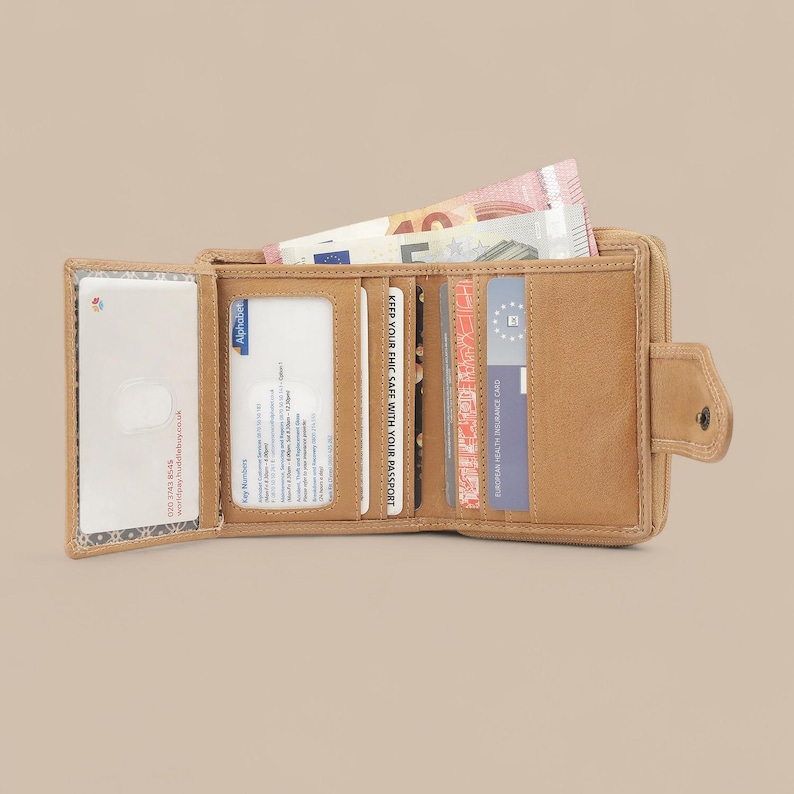 May include: A tan bi-fold wallet with a snap closure, featuring multiple interior card slots, a clear ID window, and a currency compartment containing a five-euro banknote. The wallet is open, displaying various cards and identification documents.