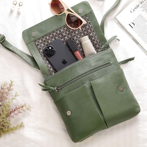 May include: Olive green leather crossbody bag with a flap closure, a front pocket, and a zipped main compartment. Inside the bag are a black smartphone, lip gloss, and a tube of shave gel. Sunglasses and earrings are also visible.